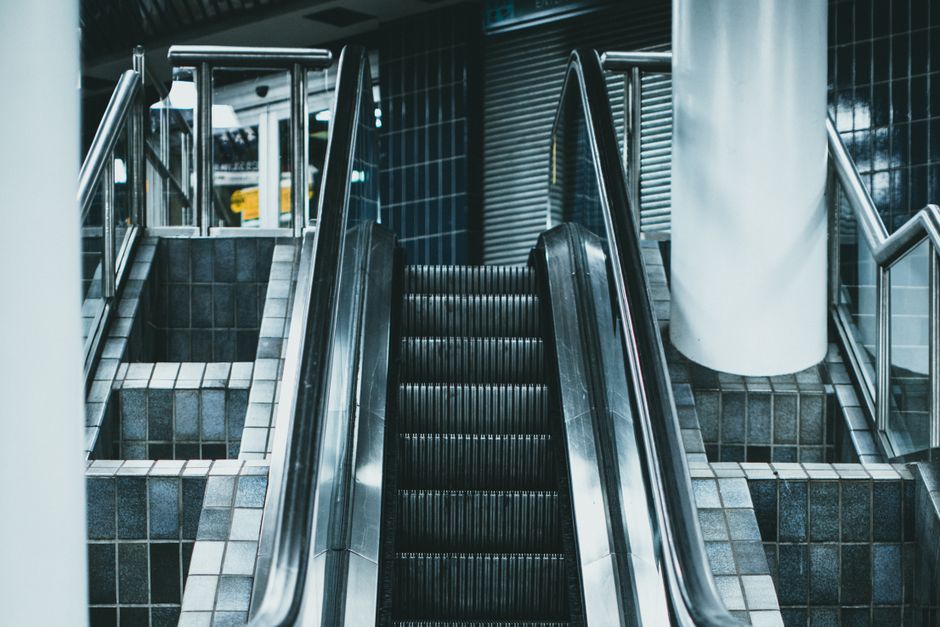 Inside a modern building near Kidbrooke Station, an escalator and a staircase are visible, both leading to upper levels. The escalator, made of metal with black steps, is centrally positioned, with its handrails on both sides. To the right of the escalator, a staircase with tiled steps and metal handrails runs parallel, descending towards the lower level. Adjacent to the staircase, a large white cylindrical column partially obscures the view. The surrounding walls are clad in dark blue tiles, creating a sleek, contemporary environment. Bright lighting illuminates the area, highlighting the metallic surfaces of the escalator and the tiled textures of the stairs. In the foreground, a section of a pavement with concrete and small gravel is visible through a doorway or open space, suggesting an interior setting with accessible passageways. This setting is indicative of a transport hub or public building where moving or housing relocation services by Man with Van Kidbrooke could be coordinated, involving careful handling of furniture and packing materials during home relocation or furniture transport processes.