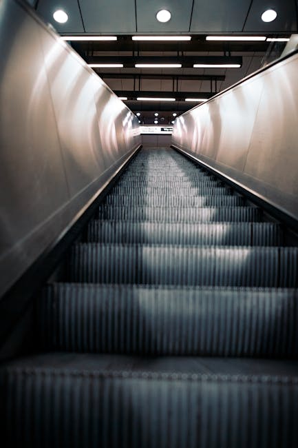A downward view of an escalator in an indoor setting, with metal steps and silver handrails on both sides, leading towards a brightly lit area with a high ceiling featuring evenly spaced ceiling lights. The escalator is situated inside a modern building, possibly an airport or train station, with a dark flooring surrounding the escalator entrance. This image is related to house removals and moving services, illustrating the transport of belongings through commercial transit hubs. The clean, professional environment aligns with the logistical aspect of furniture transport and packing during home relocation, as offered by Man with Van Kidbrooke. The scene emphasizes the movement process involved in loading or transferring items through transportation channels, with no objects or boxes visible in the image itself, but the setting naturally references large-scale moving operations.