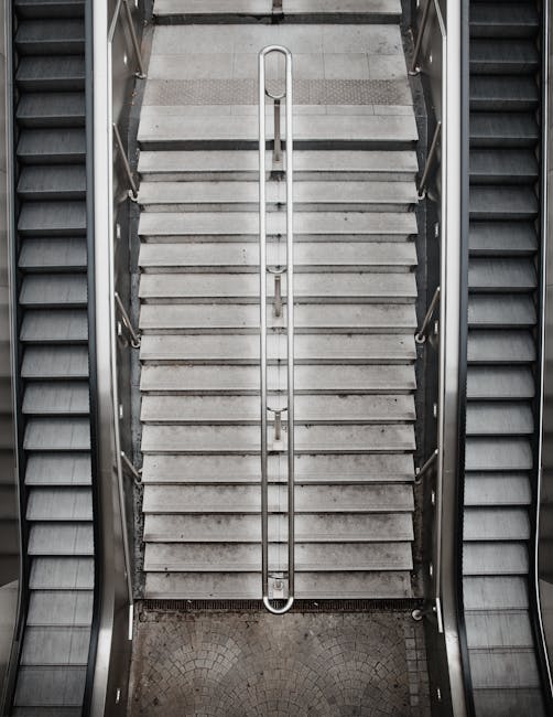 Inside a modern building near Kidbrooke Station, an escalator and a staircase are visible, both leading to upper levels. The escalator, made of metal with black steps, is centrally positioned, with its handrails on both sides. To the right of the escalator, a staircase with tiled steps and metal handrails runs parallel, descending towards the lower level. Adjacent to the staircase, a large white cylindrical column partially obscures the view. The surrounding walls are clad in dark blue tiles, creating a sleek, contemporary environment. Bright lighting illuminates the area, highlighting the metallic surfaces of the escalator and the tiled textures of the stairs. In the foreground, a section of a pavement with concrete and small gravel is visible through a doorway or open space, suggesting an interior setting with accessible passageways. This setting is indicative of a transport hub or public building where moving or housing relocation services by Man with Van Kidbrooke could be coordinated, involving careful handling of furniture and packing materials during home relocation or furniture transport processes.
