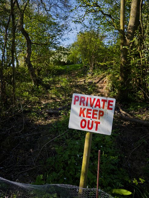 A white rectangular sign with red and orange text reading 'PRIVATE KEEP OUT' is mounted on a yellow wooden post situated at the edge of a grassy pathway leading into a wooded area. The pathway is surrounded by green trees and foliage, with some sunlight filtering through the leaves. In the background, dense greenery and tree branches create a natural environment. The scene appears outdoors, possibly indicating restricted access to a private property or area, with the sign serving as a warning. The surrounding area includes some mesh fencing visible at the bottom of the image, suggesting the boundary of the restricted zone, and the overall setting emphasizes the importance of security and privacy. This type of signage is commonly used in property management and land protection to communicate no entry without permission, aligning with home relocation considerations involving property boundaries and secure access.