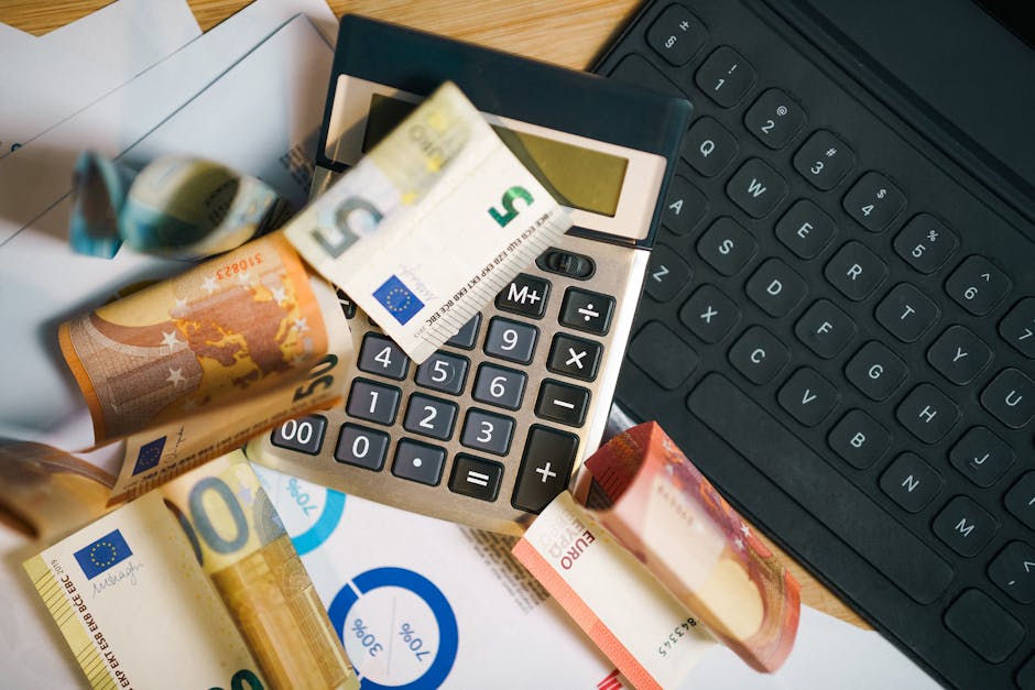 A close-up view of a home office or work desk during a house move, featuring a calculator with a five euro banknote resting on its display, alongside scattered euro notes of various denominations on the wooden surface. To the right, a black computer keyboard is partially visible, and beneath the calculator, there are sheets of white paper. The scene captures the process of packing and organizing financial or moving-related documents, with a focus on the financial aspect of a house relocation service provided by Man with Van Kidbrooke, a company specialising in removals and furniture transport within the UK.