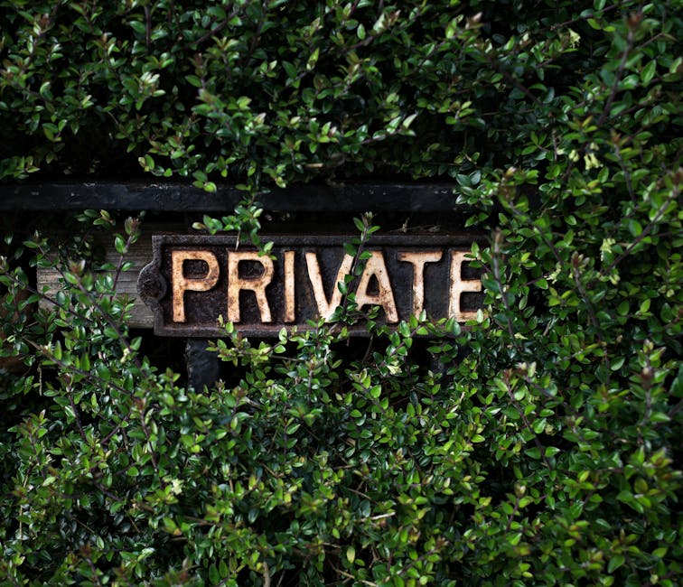 A white rectangular sign with red and orange text reading 'PRIVATE KEEP OUT' is mounted on a yellow wooden post situated at the edge of a grassy pathway leading into a wooded area. The pathway is surrounded by green trees and foliage, with some sunlight filtering through the leaves. In the background, dense greenery and tree branches create a natural environment. The scene appears outdoors, possibly indicating restricted access to a private property or area, with the sign serving as a warning. The surrounding area includes some mesh fencing visible at the bottom of the image, suggesting the boundary of the restricted zone, and the overall setting emphasizes the importance of security and privacy. This type of signage is commonly used in property management and land protection to communicate no entry without permission, aligning with home relocation considerations involving property boundaries and secure access.