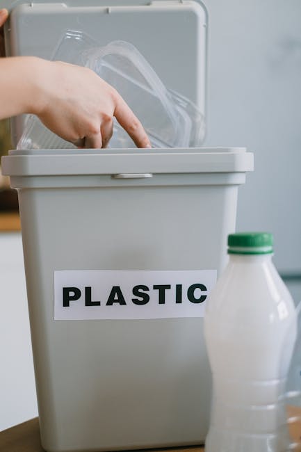 A close-up view of a grey plastic recycling bin labeled 'PLASTIC' in bold black letters, positioned on a wooden surface indoors. The bin's lid is partially open, revealing a clear plastic container inside. To the right of the bin, there is a white plastic bottle with a green cap. The background is blurred, suggesting a domestic setting. This image relates to household waste management, an essential aspect of home relocation and packing logistics, as managed by Man with Van Kidbrooke for efficient removal services.