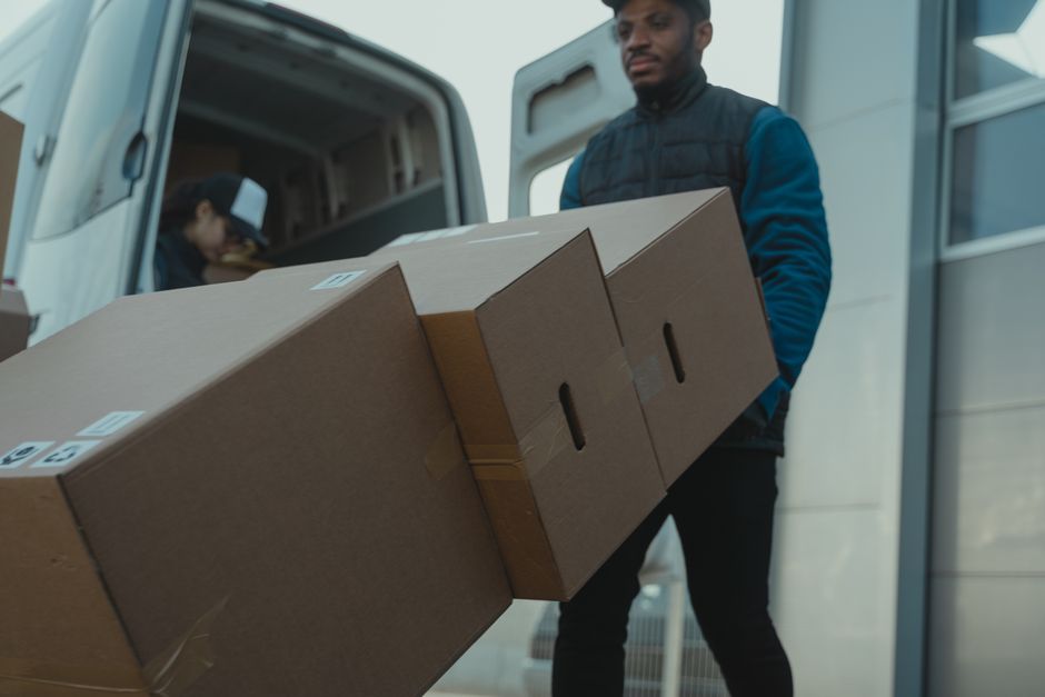A man in a dark blue jacket and black trousers is engaged in the loading process outside a property, carrying three large cardboard boxes stacked together with tape, towards a white moving van. Inside the van, a person wearing a black cap and a grey jacket is visible, handling additional packing materials or boxes. The scene takes place on a paved area adjacent to a building with a large glass door or window. The boxes are secured with packing tape, and some have labels attached, indicating organized packing and home relocation activities. The background includes a portion of the van with its open rear door, showcasing the interior lined with packaging supplies and other boxes. The environment appears well-lit, suggesting daytime, and the setup is typical of furniture transport, packing, and moving logistics coordinated by Man with Van Kidbrooke.