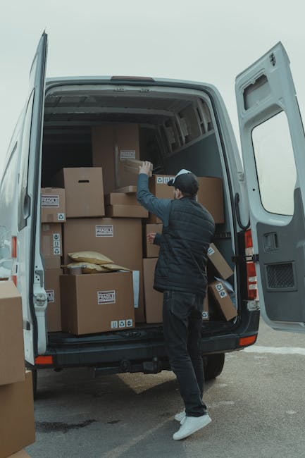 A man in a dark blue jacket and black trousers is engaged in the loading process outside a property, carrying three large cardboard boxes stacked together with tape, towards a white moving van. Inside the van, a person wearing a black cap and a grey jacket is visible, handling additional packing materials or boxes. The scene takes place on a paved area adjacent to a building with a large glass door or window. The boxes are secured with packing tape, and some have labels attached, indicating organized packing and home relocation activities. The background includes a portion of the van with its open rear door, showcasing the interior lined with packaging supplies and other boxes. The environment appears well-lit, suggesting daytime, and the setup is typical of furniture transport, packing, and moving logistics coordinated by Man with Van Kidbrooke.