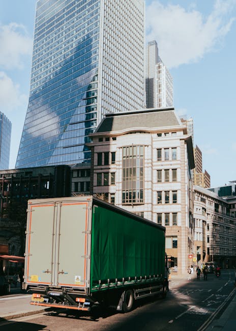 A large green cargo truck with a closed cargo area driving through a city street surrounded by tall modern office buildings with glass facades and older stone buildings. The truck is in motion along t