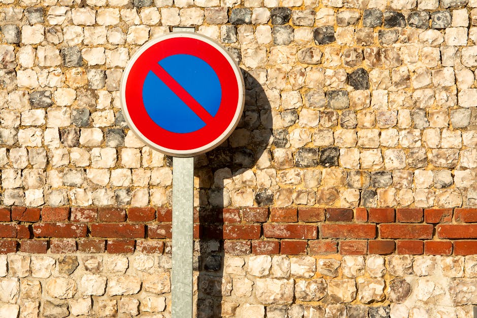 A round no parking traffic sign with a red border, blue interior, and a red diagonal line across the center, mounted on a metal pole against a textured stone wall with various shades of beige, cream, and gray, and a horizontal row of reddish-brown bricks at the bottom. The sign casts a shadow onto the wall, indicating sunlight from the right side. The setting suggests a street or urban area where temporary parking restrictions may impact home relocation or furniture transport, as handled by Man with Van Kidbrooke, especially during moving or packing and moving processes involving loading or unloading near the property.