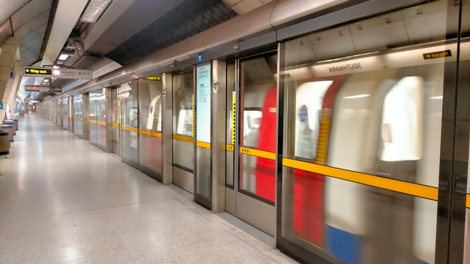 A long, empty underground train station platform with a row of glass sliding doors separating the waiting area from the train tracks. The platform features a clean, polished stone floor, and the doors have yellow safety lines near their midsection. Behind the glass, a train with a white exterior and red and blue accents is visible, with its interior faintly showing through the windows. Overhead lighting illuminates the area evenly, and signs indicating way out and other directions are mounted above the platform. This station setting relates to house removals and moving logistics, highlighting the importance of transportation and planning during home relocations, as managed by services like Man with Van Kidbrooke.