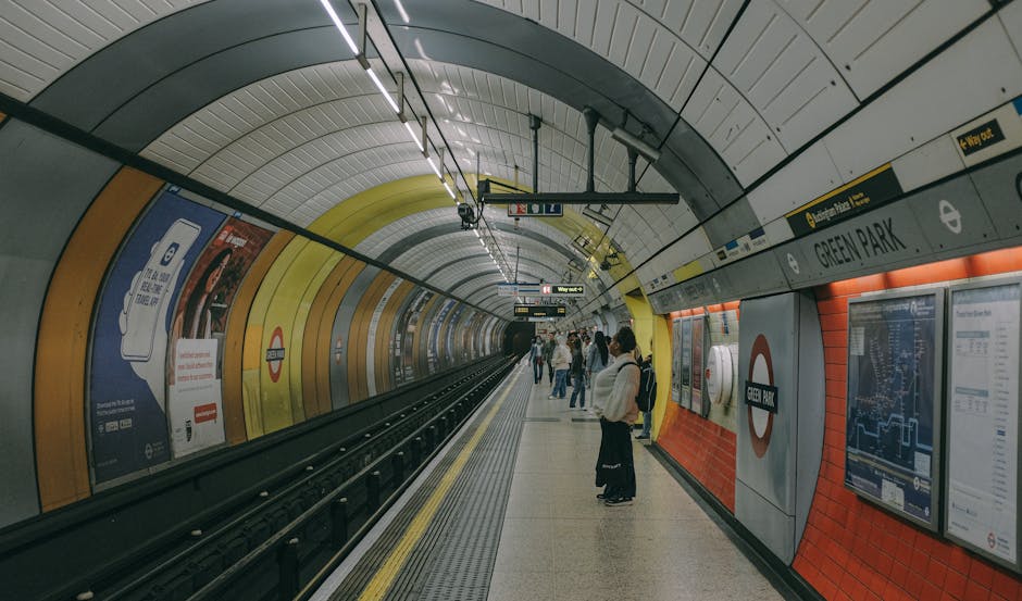 A long, empty underground train station platform with a row of glass sliding doors separating the waiting area from the train tracks. The platform features a clean, polished stone floor, and the doors have yellow safety lines near their midsection. Behind the glass, a train with a white exterior and red and blue accents is visible, with its interior faintly showing through the windows. Overhead lighting illuminates the area evenly, and signs indicating way out and other directions are mounted above the platform. This station setting relates to house removals and moving logistics, highlighting the importance of transportation and planning during home relocations, as managed by services like Man with Van Kidbrooke.